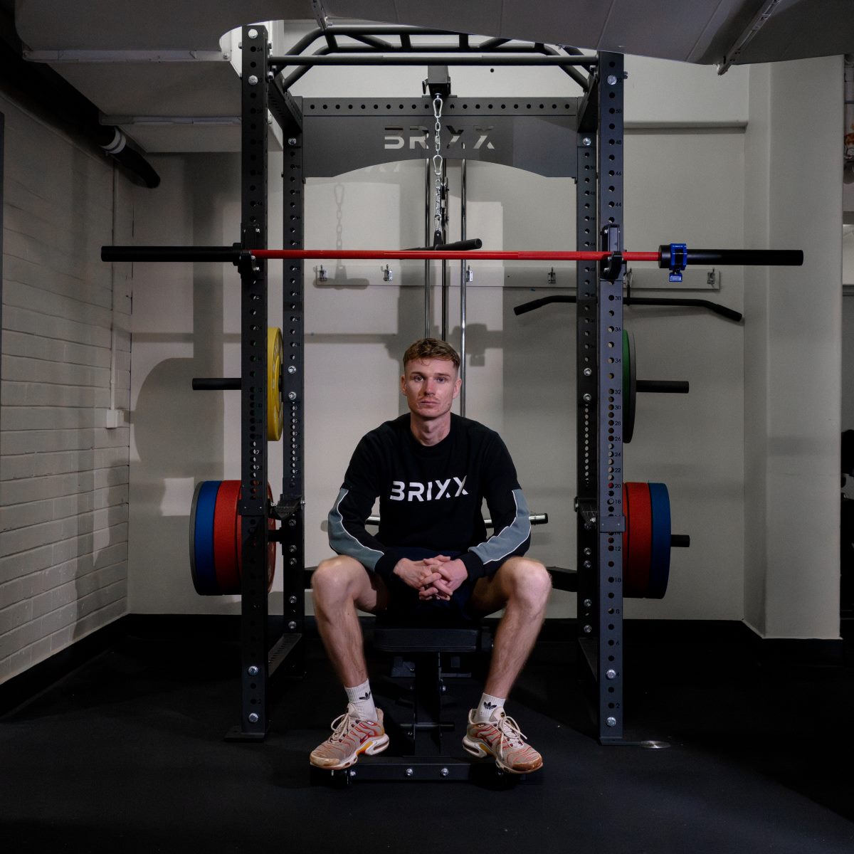 Man sitting on an adjustable bench inside a BRIXX power rack with lat pulley, barbell and colour bumper plates